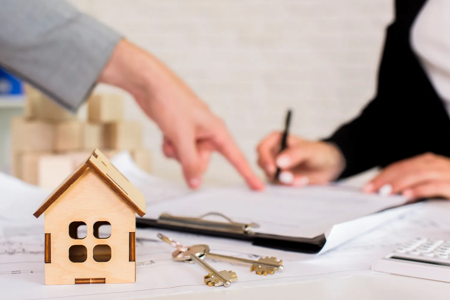 A miniature wooden house near keys and documents, as a hand gestures to a contract being signed by another person